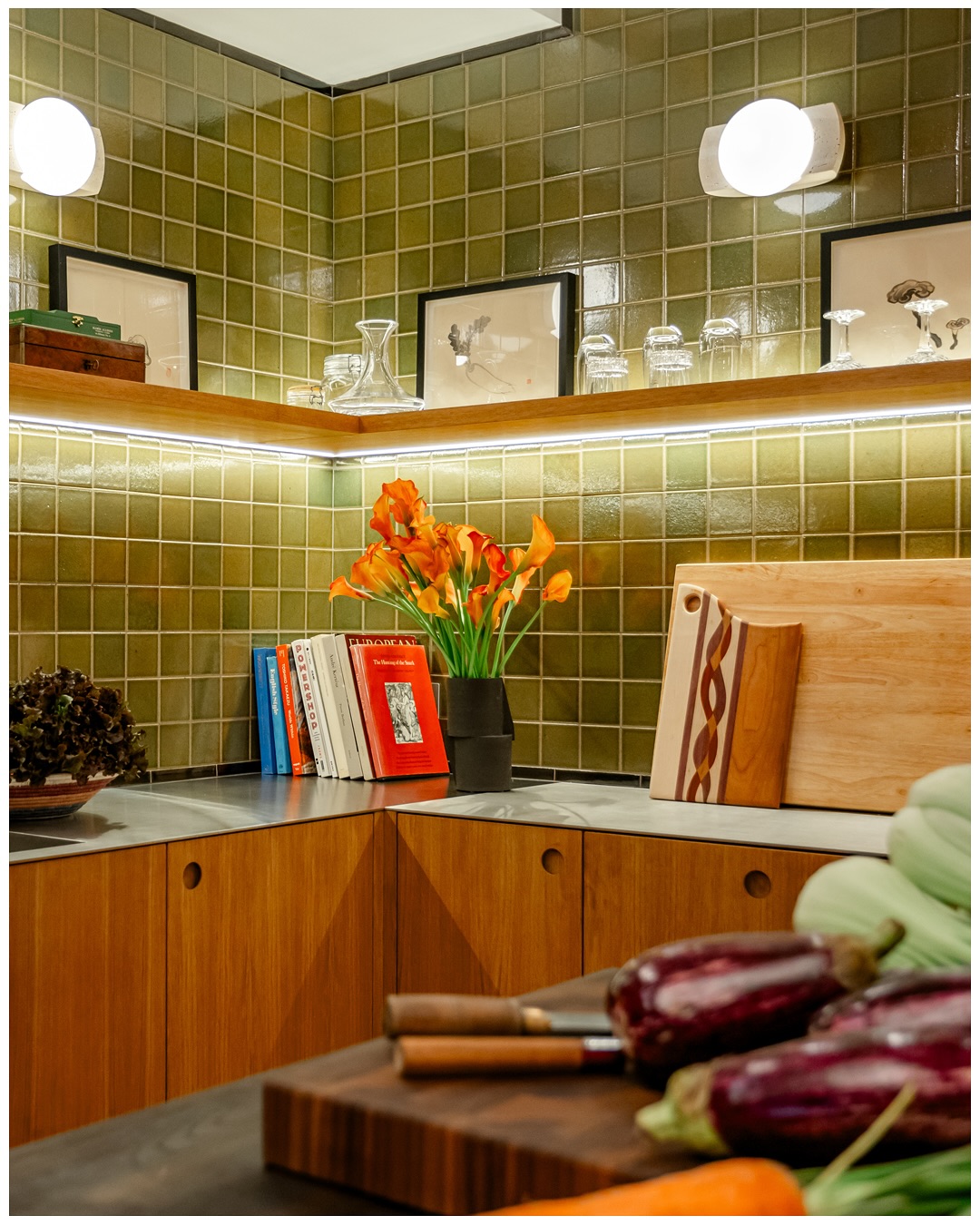 Corner counter with a floating wood shelf, flowers, and cookbooks against green tile